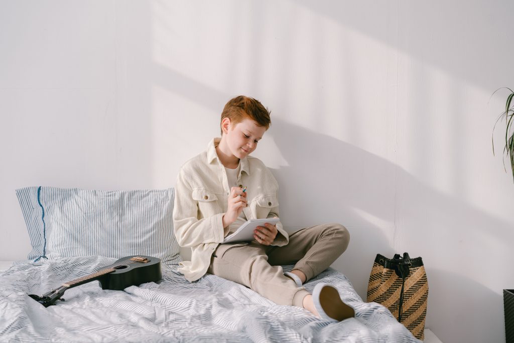 Tiener schrijft in een notitieboek op zijn bed, met een gitaar ernaast in een lichte tienerkamer